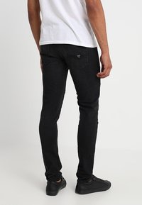 Jeans de mezclilla negros con corte ajustado, que cuentan con un sutil logo en el bolsillo trasero y un material texturizado. Combinados con zapatillas negras.