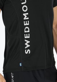 Sort atletisk skjorte med "SVERIGE" i fed hvid tekst på siden. Inkluderer et lille svensk flag ved bunden. Lavet af glat stof.