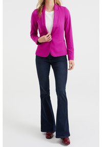 Blazer ajusté fuchsia avec un bouton unique, des manches longues et des revers crantés, associé à un jean foncé évasé et des chaussures vernies rouge foncé.