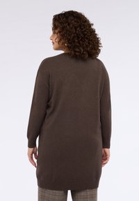Maglione lungo marrone con vestibilità ampia, orlo a coste e maniche lunghe, abbinato a pantaloni a quadri. La texture sembra morbida e lavorata a maglia.