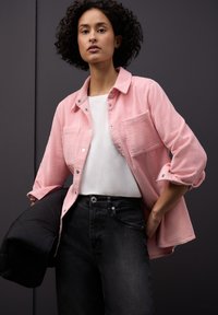 Różowa koszula sztruksowa z kołnierzykiem zapinanym na guziki, z kieszeniami z przodu, noszona na białym t-shircie i zestawiona z czarnymi dżinsami.