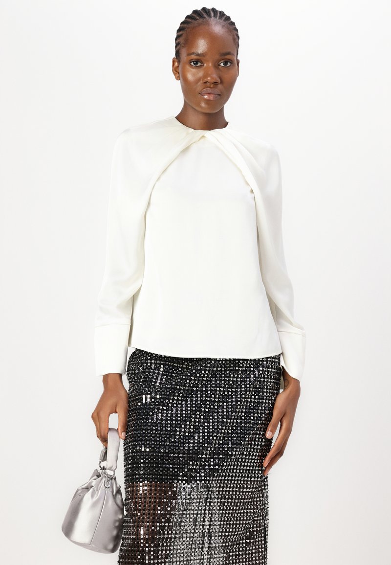Biała bluzka z długim rękawem z detalem w postaci węzła przy szyi, w połączeniu z czarną spódnicą ołówkową wykonaną z cekinów. Modelka trzyma małą srebrną torebkę.