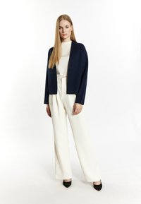 Navy cropped cardigan over een witte coltrui, gecombineerd met hoge witte broek met wijde pijpen en zwarte puntige hakken.