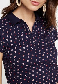 Marinblå polotröja i lätt tyg, med ett litet hjärt-mönster i röd och vit, samt en två-knapps knappslå vid kragen.