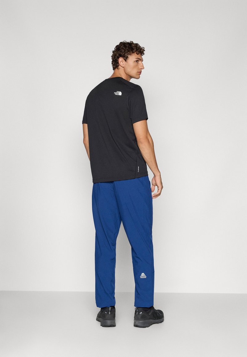 Schwarzes Kurzarmhemd mit Logo auf dem Rücken, kombiniert mit blauen Hosen, die ein Logo am Unterschenkel haben, und schwarzen Schuhen.