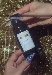 Black, squeezable bottle labeled "Glass OIl" by Pat McGrath Labs. Hands feature long, sparkly nails with multi-colored rhinestones. Gold sequined background.
