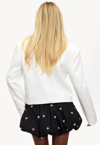 Blazer structuré blanc à manches longues, associé à un skort noir orné de motifs floraux blancs. Design épuré et texture lisse.