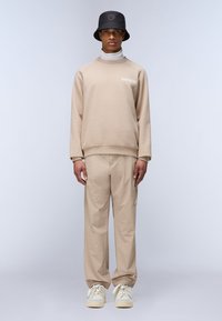 Sudadera beige con puños acanalados, combinada con pantalones beige a juego que tienen tobillos elásticos, llevados con zapatillas blancas y un sombrero de cubo negro.