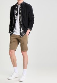 Black bomber jacket over a patterned shirt, white t-shirt, tan shorts, and white sneakers with visible laces and textured socks.