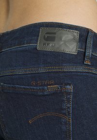 Jeans de mezclilla oscura con un parche de cuero de G-Star RAW, costuras naranjas y un pequeño logo metálico de G-Star cerca del bolsillo.
