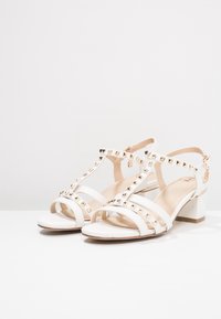 Valkoiset sandaalit, joissa on paksu korko, sisältävät useita hihnoja, kultaisia niittejä ja säädettävän nilkkahihnan. Sileä pinta ja minimalistinen muotoilu.