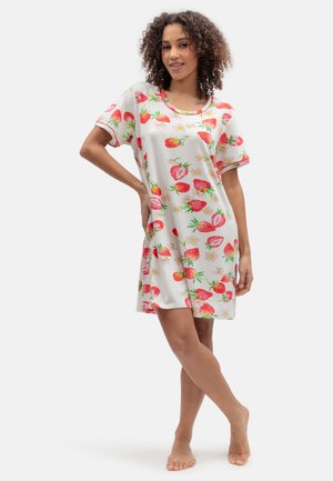 Vrouw staat blootsvoets, draagt een korte witte jurk met een rood aardbeien- en bloemenprint, glimlacht met één hand op haar heup.