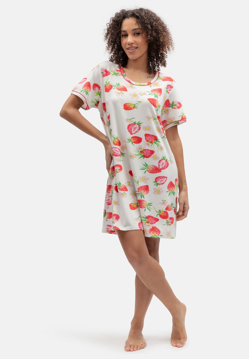 Kobieta stojąca boso, ubrana w krótką białą sukienkę w czerwone truskawki i kwiaty, uśmiechająca się z jedną ręką na biodrze.