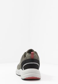 Zapatilla deportiva con parte superior de malla en verde oliva, acentos negros y una pestaña en el talón roja. Cuenta con una suela acolchada con una banda de rodadura de goma en contraste.