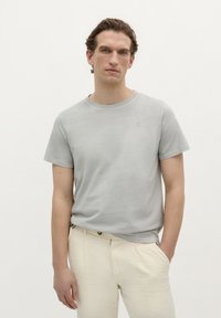 Lichtgrijs T-shirt met korte mouwen gemaakt van zachte stof. Het heeft een ronde hals en een subtiel logo op de linkerborst. Gecombineerd met crèmekleurige broek.