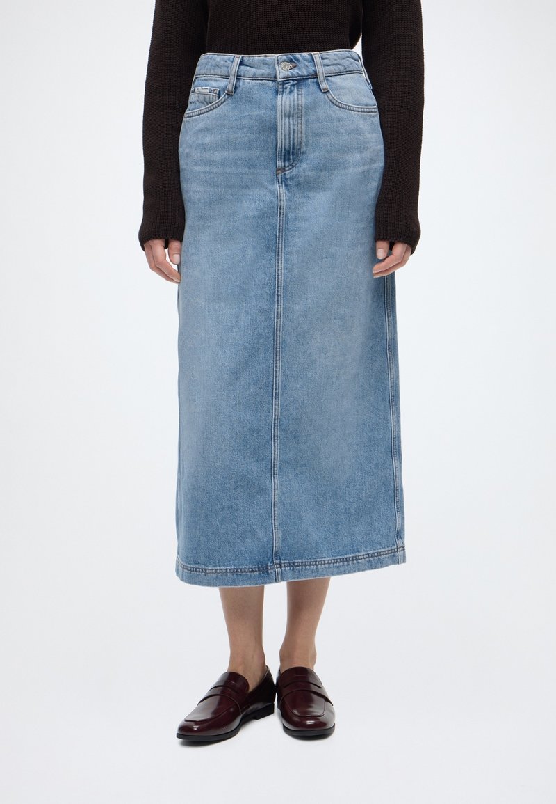 Άτομο που φοράει μίντι ανοιχτό μπλε τζιν φούστα, σκούρο μακρυμάνικο τοπ και σκούρα κόκκινα μοκασίνια, στέκεται μπροστά σε απλό λευκό φόντο.