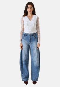 Weißes Spitzenshirt mit langen Ärmeln und V-Ausschnitt, kombiniert mit lockeren blauen Jeans. Die Schuhe sind dunkelburgunderrot, und das Modell trägt minimalen Schmuck.