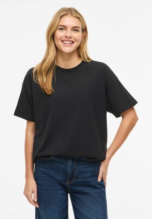 Femme souriante aux longs cheveux blonds portant un t-shirt noir ample à manches courtes et un jean bleu, debout avec une main sur la hanche.