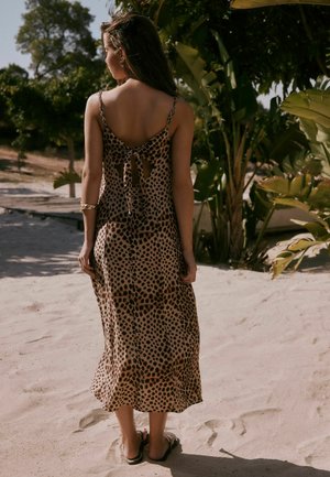 Kobieta ubrana w brązowo-czarną sukienkę bez rękawów w cętki z kokardą z tyłu, stojąca boso na piaszczystym podłożu w pobliżu zielonych roślin.