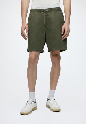 Homme portant un short vert olive à cordon, des chaussettes blanches et des baskets blanches, debout devant un fond clair uni.