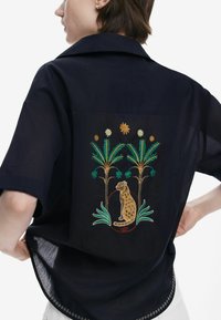 Lühikeste varrukatega tumesinine särk, mille seljal on erksate värvidega brodeeritud leopard ja palmipuud. Diskreetne tekstuuri detail.