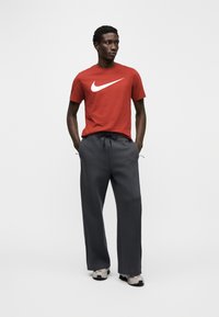 Tricou roșu din bumbac cu logo-ul Nike alb, asortat cu pantaloni sport gri închis cu talie cu șnur și adidași gri deschis.