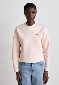 Svijetlosmeđi dukserica s raglan rukavima, rebrastim okovratnikom i zelenim Lacoste logotipom na prsima, uparen s svijetloplavim trapericama.