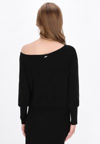 Pulover negru, cu umeri descoperiți, cu mâneci lungi, manșete din bumbac și o textură netedă. Prezintă un mic eticheta argintie pe spate.