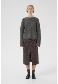 Pull en maille gris et fluffy à manches longues associé à une jupe en cuir marron avec une fente à l'avant. Le modèle porte des chaussettes grises et des chaussures noires.
