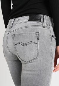 Persona que lleva vaqueros de un gris claro con detalle bordado en el bolsillo trasero y una camiseta negra, mostrando la vista trasera desde la cintura hasta la mitad del muslo.