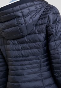 Marineblå pufferjakke med quiltet tekstur og hætte. Har et slankt design, letvægtsmateriale og en tætsiddende silhuet.