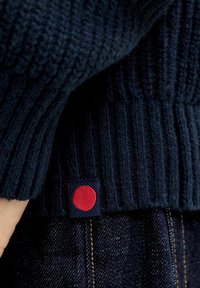 Detail eines marineblauen gestrickten Pullovers mit geripptem Muster und einem roten, runden Etikett, das auf einen dunklen Denim-Hintergrund aufgenäht ist.