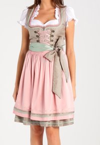 Femme portant une robe traditionnelle dirndl rose et beige avec une blouse blanche, un corsage lacé et un grand nœud noué à la taille.