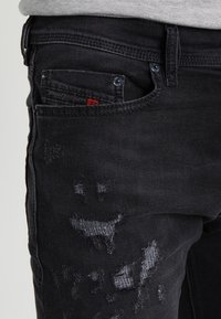Jeans de mezclilla negra con parches desgastados en los muslos y un pequeño bolsillo frontal etiquetado "Diesel Industry."