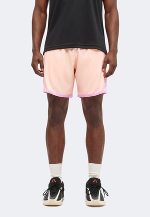 Person trägt rosa Sportshorts mit lila Saum, schwarzes Shirt, weiße Socken und schwarz-beige Laufschuhe und steht mit auseinander stehenden Füßen.