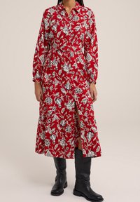 Röd blommig klänning med vita mönster, långa ärmar, knappkrage och en slits framtill. Matchas med knähöga svarta stövlar.