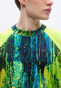 Πολύχρωμο φούτερ με ζωηρό σχέδιο πιτσιλιές σε πράσινο, μπλε και μαύρο, με μανίκια ραγκλάν νέον κίτρινου και κεντημένο λογότυπο της μάρκας.