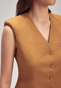 Vestido mostaza sin mangas con un profundo escote en V, tres botones forrados de tela y un bolsillo lateral; fabricado en un tejido texturizado y tejido.