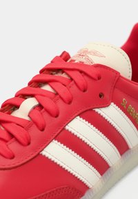 Sapatilhas vermelhas de couro com riscas brancas, atacadores vermelhos, língua bege com um logótipo e "SL BENFICA" em letras douradas na lateral.