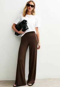 Vrouw met golvend haar draagt een wit T-shirt en een bruine gestreepte wijde broek, houdt een zwarte clutch vast en staat tegen een lichte muur.