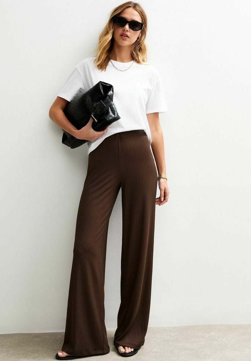 Vrouw met golvend haar draagt een wit T-shirt en een bruine gestreepte wijde broek, houdt een zwarte clutch vast en staat tegen een lichte muur.