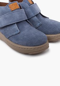 Scarpe in suede blu con cinturini in Velcro, dotate di una toppa logo marrone. La punta arrotondata e le suole in gomma beige conferiscono un design casual.