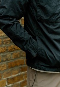 Jachetă neagră cu o croială relaxată, având un buzunar frontal, mâneci lungi și o textură fină, purtată peste pantaloni de culoare deschisă.