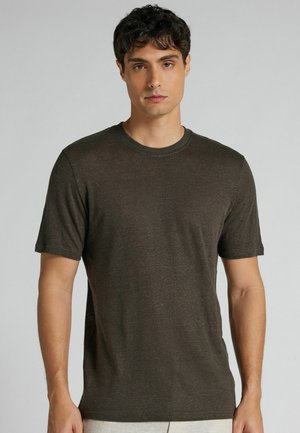 Hombre con pelo corto y oscuro que lleva una camiseta de manga corta de cuello redondo lisa de color verde oliva oscuro sobre un fondo gris claro.