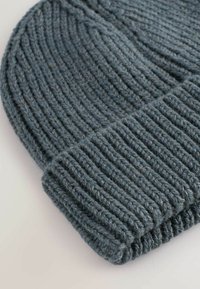 Căciulă tricotată gri, confecționată din lână groasă, cu o textură riburoasă și margine întoarsă. Moale și călduroasă, cu o croială strânsă.