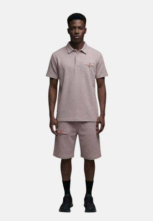 Homme portant un polo à manches courtes texturé assorti et un short arrivant aux genoux avec des chaussettes et chaussures noires, debout devant un fond uni.