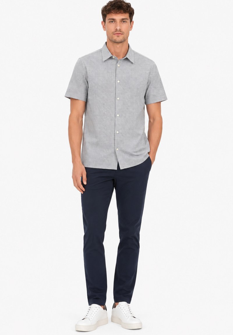 Homme debout portant une chemise grise à motifs à manches courtes boutonnée, un pantalon bleu foncé, des baskets blanches, avec une main dans la poche sur fond blanc.