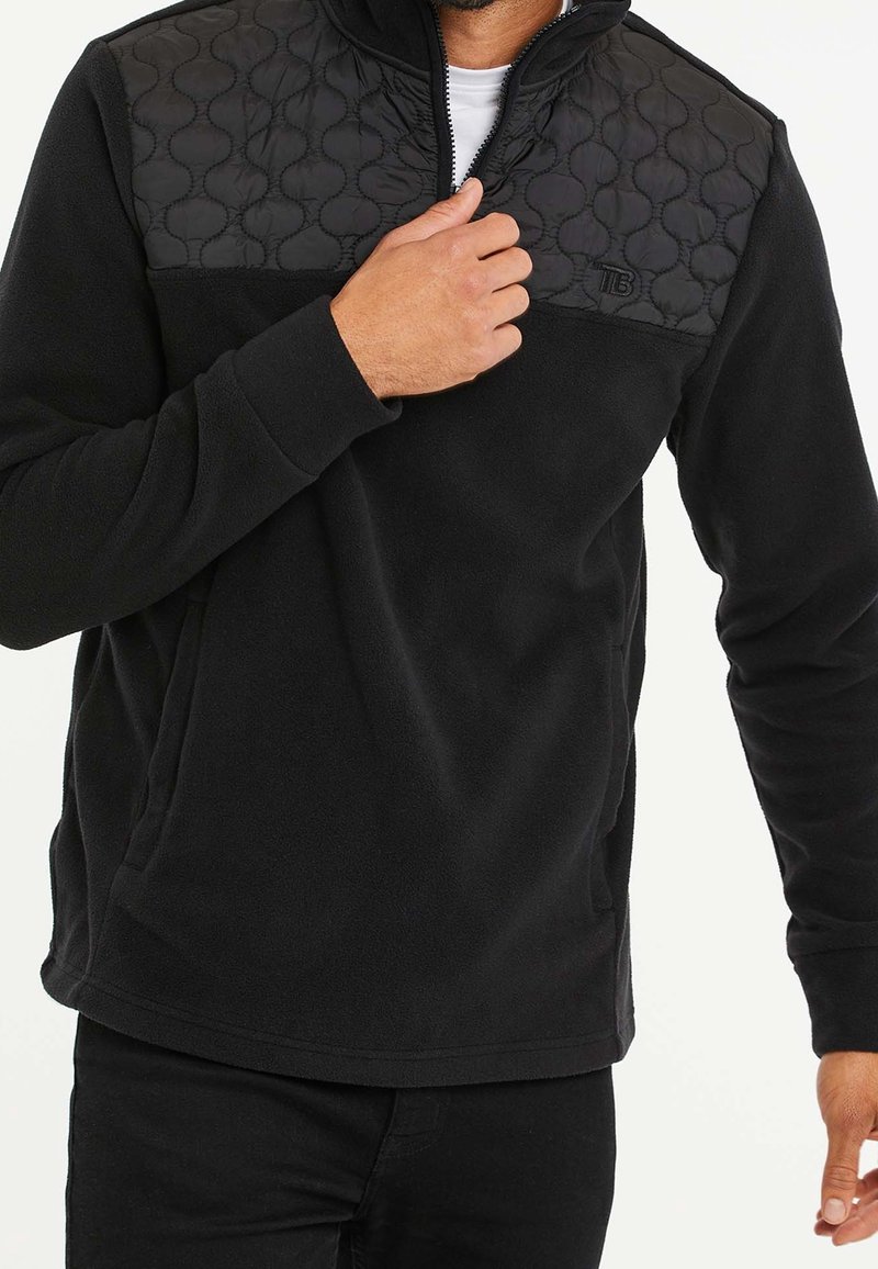 Homme portant un pull en polaire noir avec un panneau matelassé sur la poitrine et une demi-fermeture éclair, ajustant le col avec la main droite.