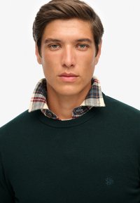 Suéter verde oscuro con acabado texturizado, superpuesto a una camisa de cuadros en colores beige, azul marino y burdeos. Detalle sutil del logo en el suéter.
