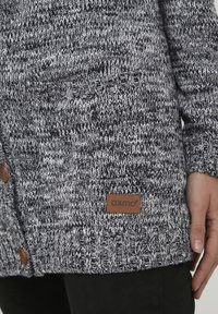 Cardigan tricoté gris et blanc avec un motif texturé, doté de boutons en bois et d'une étiquette en cuir brun en bas à l'avant.